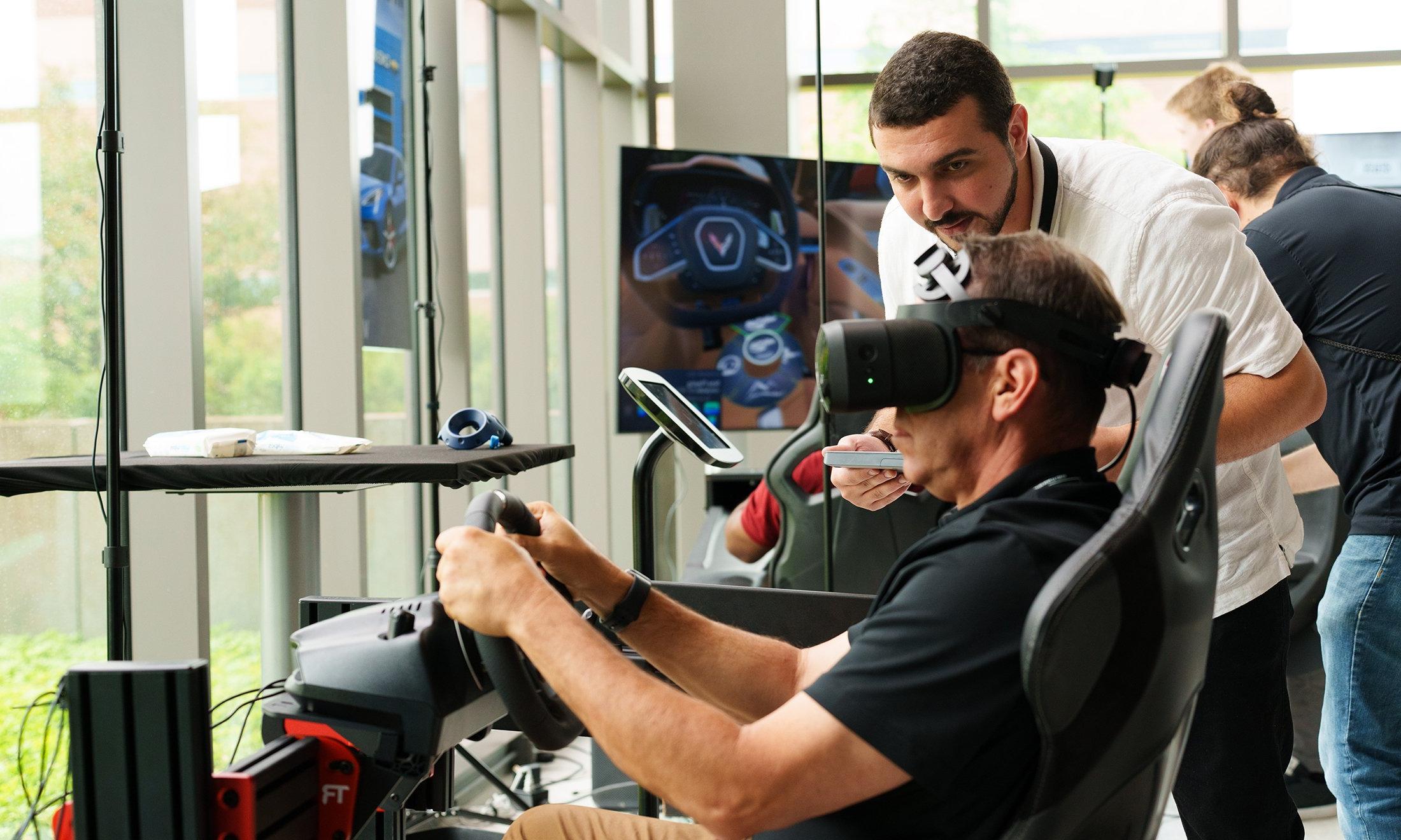 A person watching someone wearing a VR headset and using a steering wheel.